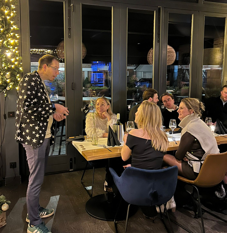 Goochelaar Tim Wijst in colbert met sterren treedt op voor een gezelschap aan een gedekte tafel tijdens een kerstborrel in een restaurant