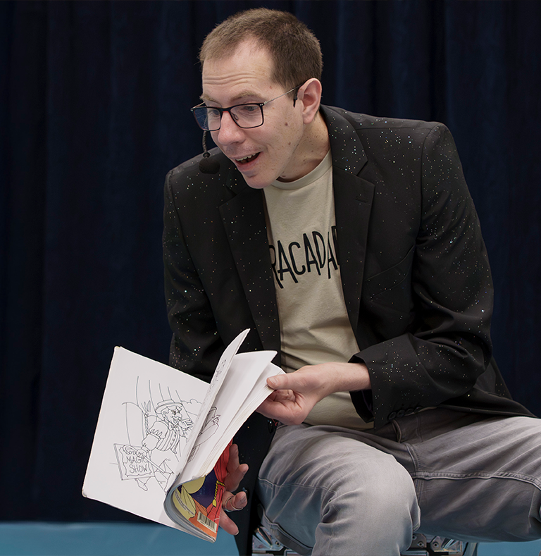 Goochelaar Tim Wijst in ABRACADABRA shirt en donker colbert met sterretjes, leest voor uit een boek tijdens een kinderboekenweek act