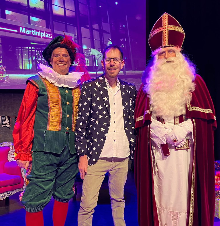 Goochelaar Tim Wijst in colbert met sterren poseert tussen Sinterklaas en een Piet op een podium in Martiniplaza